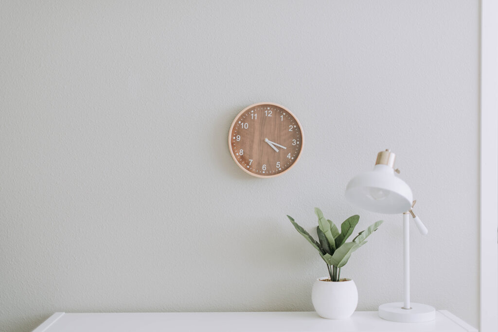 Minimalistischer Design-Arbeitsplatz mit einer hölzernen Wanduhr, die 3:53 Uhr anzeigt, einer weißen Schreibtischlampe und einer kleinen Topfpflanze auf einem weißen Schreibtisch - perfekt für Ihre praktischen Aufgaben - vor einer hellgrauen Wand.