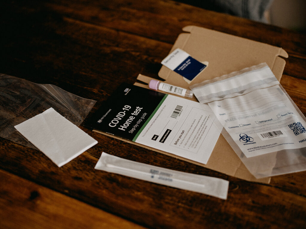 Auf einem Holztisch liegt ein COVID-19-Heimtest-Kit mit Anleitung, Tupfer, Probenbeutel und anderem Verpackungsmaterial aus der Marken-Niederlassung in München bereit.