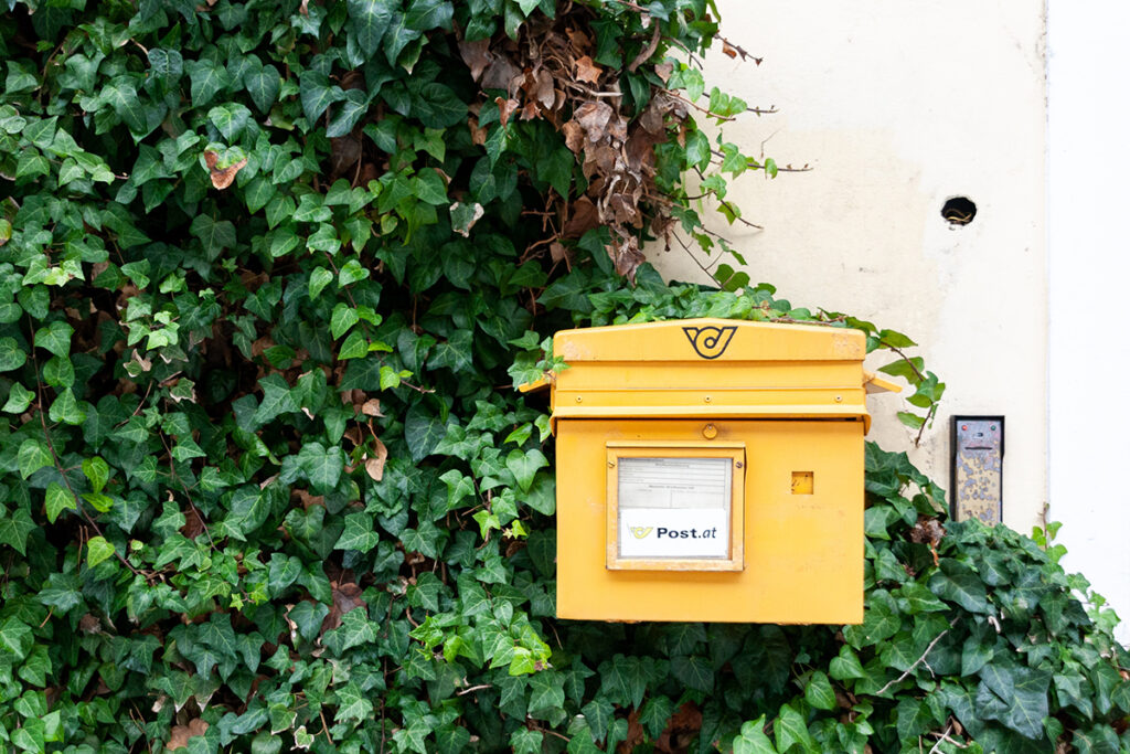Ein leuchtend gelber Briefkasten, möglicherweise von Marken wie IDEENHAUS in Nürnberg entworfen, ist an einer mit grünen Efeublättern bewachsenen Wand angebracht. Der Briefkasten hat ein Logo und ein Etikett mit der Aufschrift Post.at und steht neben einer schwarzen Türklingel oder Sprechanlage.