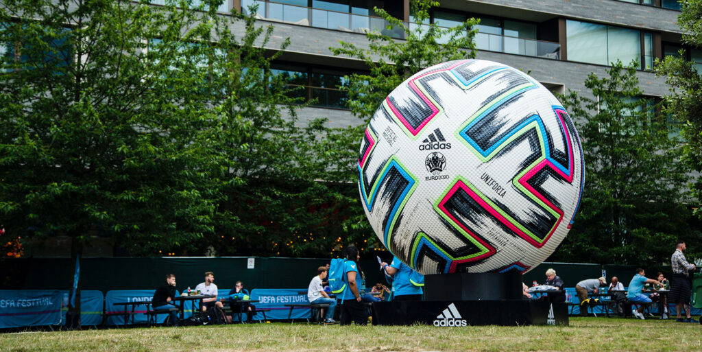 Eine riesige Fußballskulptur der UEFA Euro 2020 von Adidas ist im Freien in der Nähe von Bäumen und einem modernen Gebäude ausgestellt, während im Hintergrund Menschen an Tischen sitzen.
