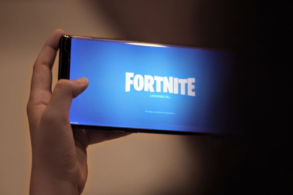 Eine Person hält ein Apple-Smartphone in der Hand, auf dem der Ladebildschirm des Spiels Fortnite angezeigt wird, wobei der Titel des Spiels auf blauem Hintergrund deutlich zu erkennen ist.