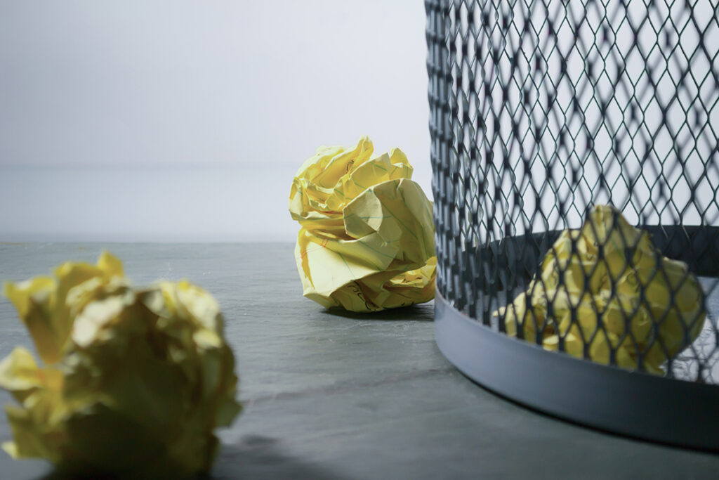 Auf einer grauen Fläche steht ein Papierkorb aus Metallgeflecht, vor dem drei zerknüllte gelbe Papiere liegen - eine subtile Anspielung auf den kreativen Prozess bei IDEENHAUS, wo Marken und Ideen geformt werden. Der Hintergrund ist schlicht und sanft beleuchtet.