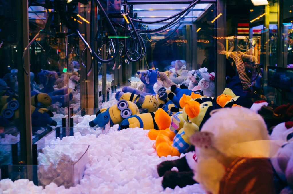 Ein hell erleuchteter Krallenautomat auf der Spielwarenmesse, gefüllt mit bunten Plüschtieren - darunter gelbe Minions, Enten und mehr - fängt die Begeisterung der Menschen ein, die sich in den Glasseiten des Automaten spiegeln.