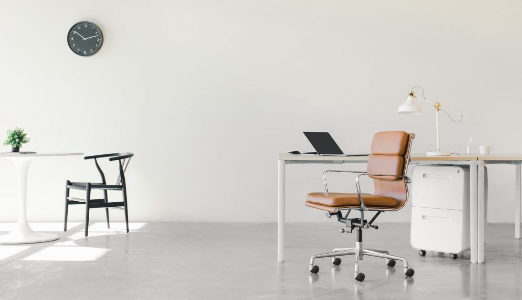 Minimalistisches Büro, entworfen für Fachkräfte, mit einem braunen Lederstuhl an einem Schreibtisch mit Laptop, Lampe und Aktenschrank. Im Hintergrund steht ein kleiner runder Tisch mit zwei schwarzen Stühlen unter einer Wanduhr vor weißen Wänden und Böden.