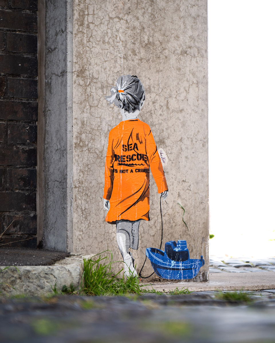 Street Art eines Kindes in einem orangefarbenen Mantel mit der Aufschrift SEA RESCUE IS NOT A CRIME auf dem Rücken, das ein blaues Spielzeugboot - Teil einer Kampagne - in der Hand hält, gemalt auf eine Betonmauer neben einem Rasenstück.