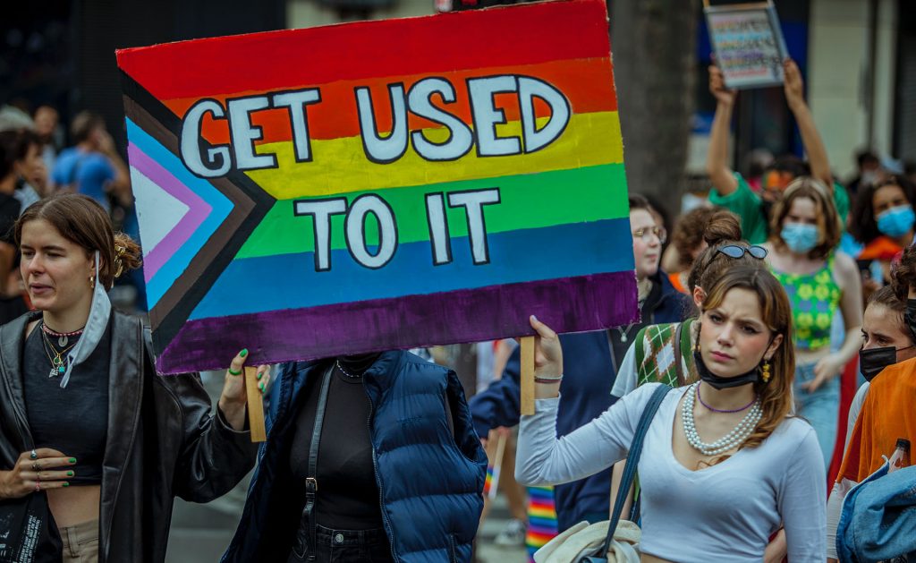 Menschen bei einer Pride-Parade halten ein buntes Schild mit Regenbogen- und Progress-Pride-Flaggenfarben, auf dem steht: "GET USED TO IT". In der Menge ist die Aktivistin Tessa Ganserer zu sehen, die mit Menschen unterschiedlichen Alters und unterschiedlicher Stile feiert.