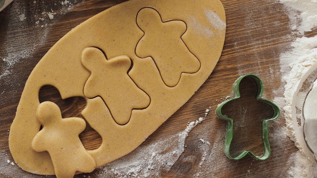 Auf einer bemehlten Holzfläche liegt ein ausgerolltes Blatt Plätzchenteig, aus dem drei Lebkuchenplätzchen ausgestochen werden, daneben liegt eine Ausstechform aus Metall. Um den Teig, die Plätzchenformen und die Ausstechform wird Mehl gestreut.