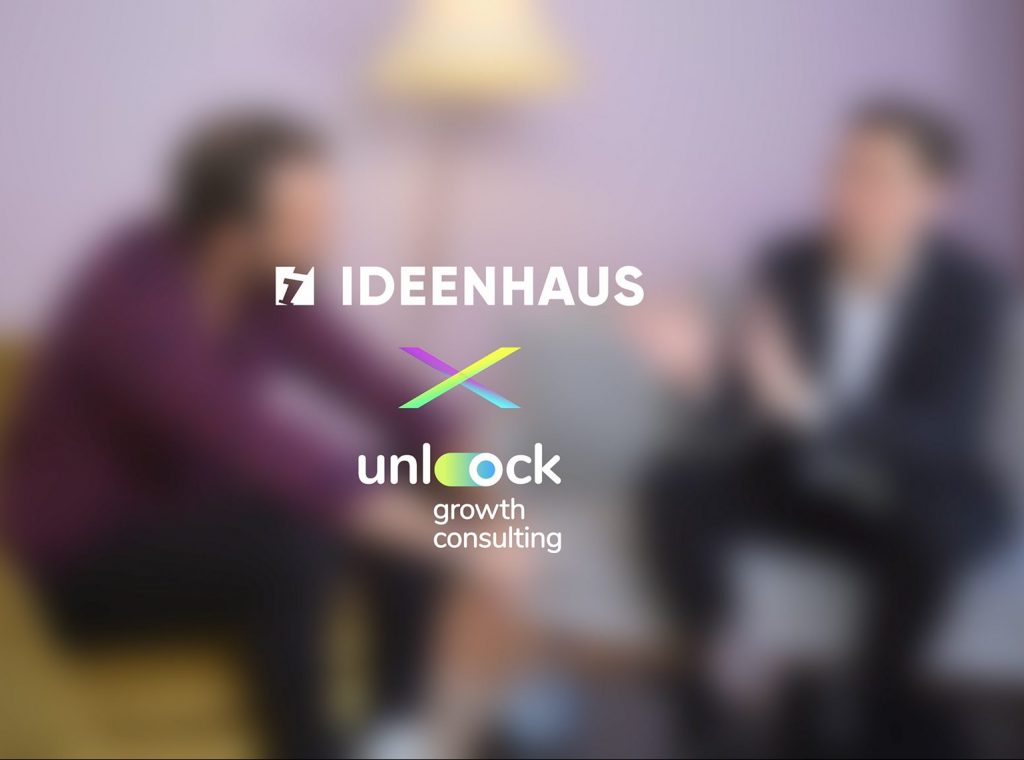 Zwei Personen sitzen und unterhalten sich in einer lockeren Umgebung mit verschwommenen Gesichtern. Die Logos von Unlock Growth und IDEENHAUS sind überlagert, während eine Lampe und ein lila Hintergrund die Szene vervollständigen.