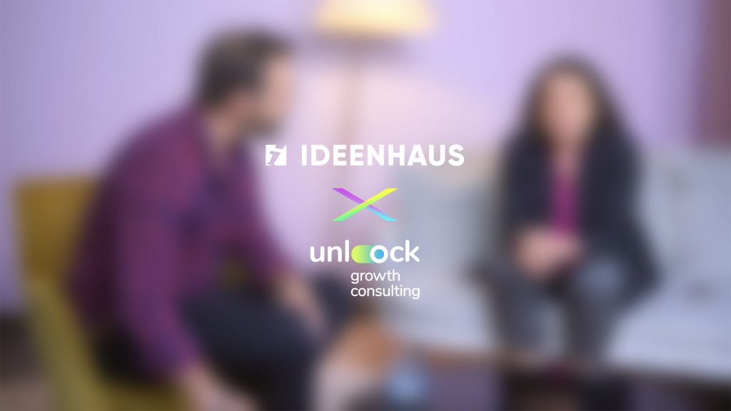 Zwei Personen sitzen sich in einem unscharfen Raum gegenüber, während in der Bildmitte die Logos von IDEENHAUS und Unlock Growth scharf eingeblendet sind.