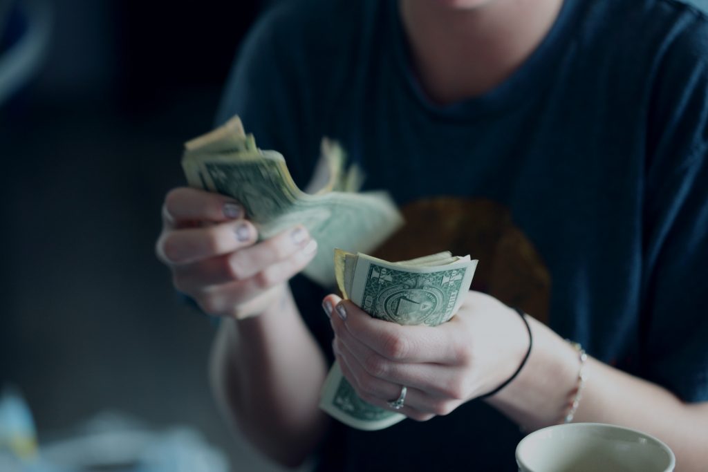 Eine Person in einem blauen Hemd zählt einen Stapel Ein-Dollar-Scheine und denkt über die Positionierung des Arbeitsgebers nach; ihr Gesicht ist unscharf, und in der unteren rechten Ecke erscheint eine weiße Tasse.