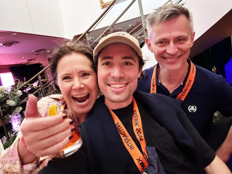 Drei lächelnde Menschen posieren auf der SXSW in einem Innenraum für ein Selfie. Die Frau links hält ein Getränk in der Hand und zeigt den Daumen nach oben, während die beiden Männer mit orangefarbenen Schlüsselbändern strahlend in die Kamera lächeln.