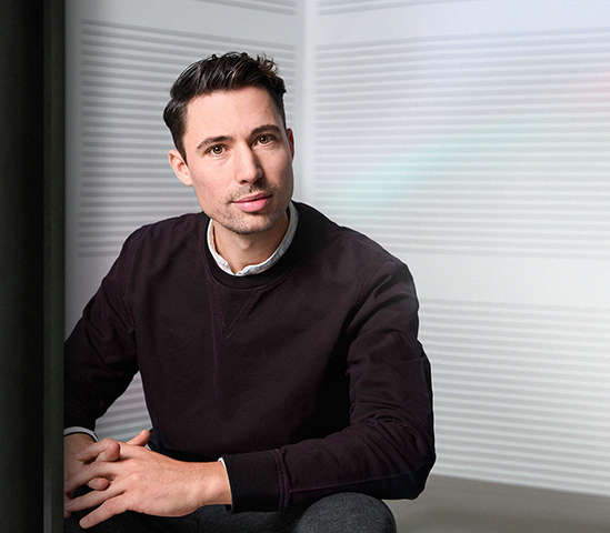 A man with short dark hair wearing a dark sweatshirt sits indoors in front of a light-colored wall with horizontal lines, looking at the camera with a neutral expression and hands clasped, pondering if AI is the gravedigger of creativity.