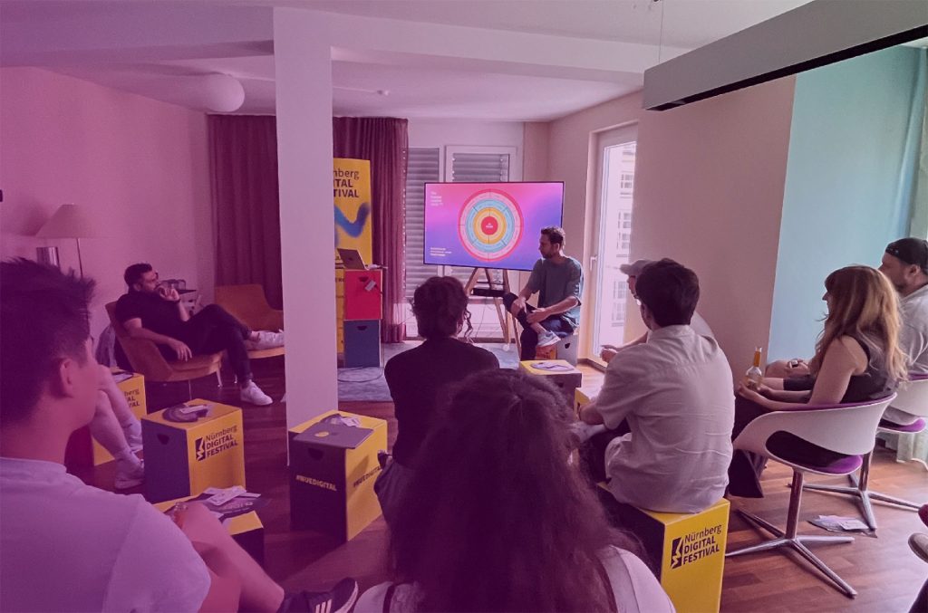 Eine Gruppe von Menschen sitzt in einem Wohnzimmer auf Stühlen und gelben Kästen und hört einem Moderator zu, der eine bunte kreisförmige Grafik über Künstliche Intelligenz auf einem Bildschirm anzeigt. Im Hintergrund sind leuchtende Banner des Nürnberg Digital Festivals zu sehen.