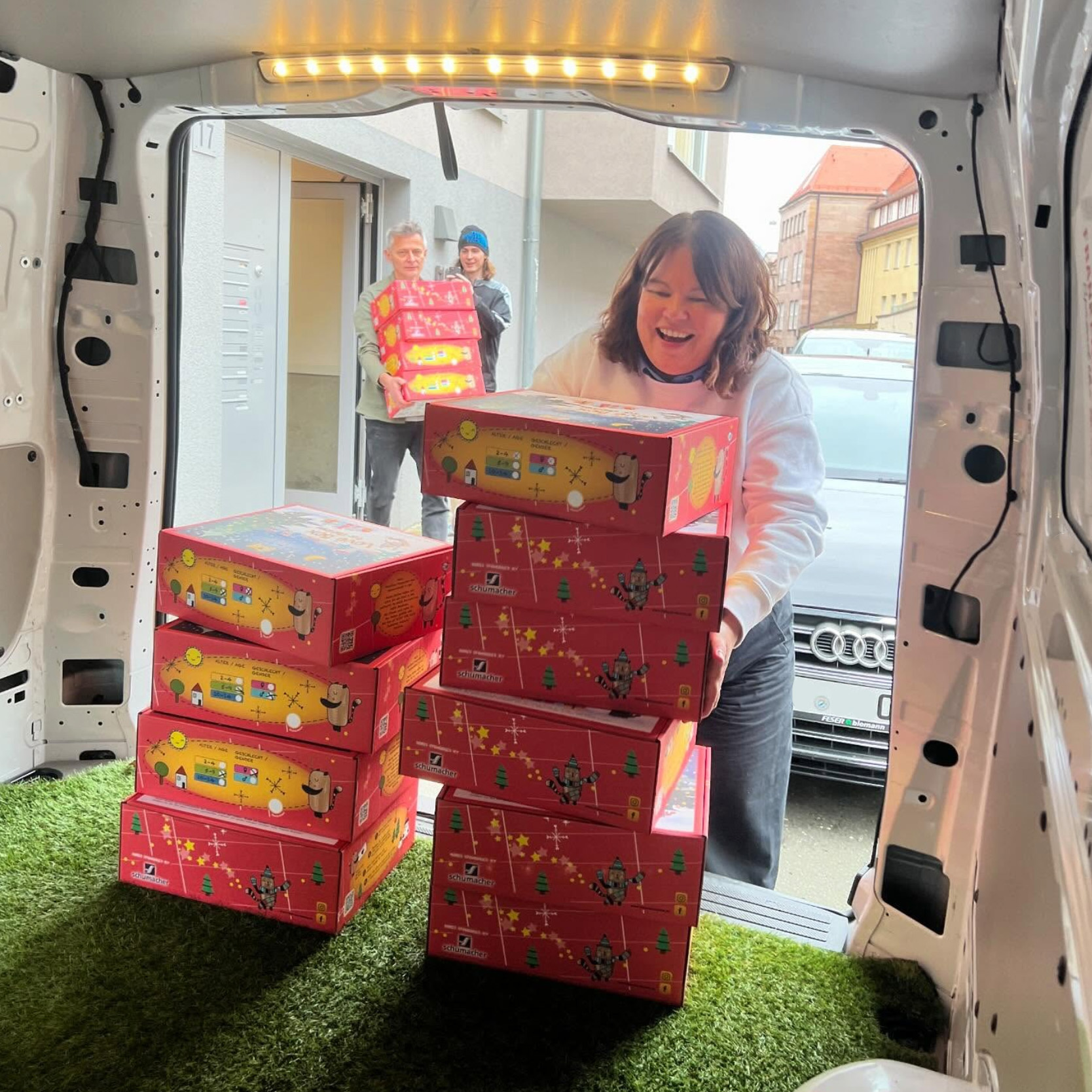 Eine lächelnde Frau lädt festliche rote Kartons in einen Lieferwagen mit grünem Teppichboden, während zwei Personen im Hintergrund weitere Kartons auf einer Straße in der Stadt zum Lieferwagen tragen.