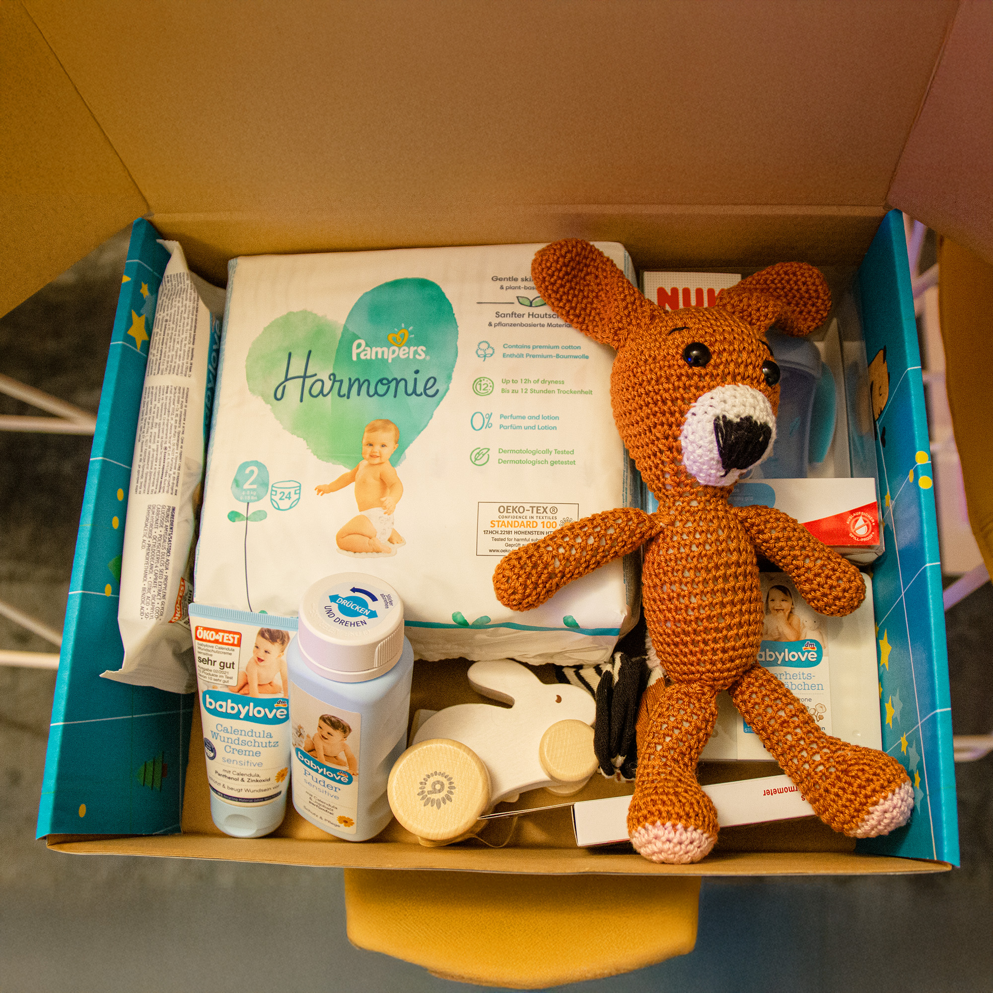 Ein Karton enthält Babyartikel wie Pampers-Windeln, Lotion, Puder, Creme, ein Holzspielzeug, einen Schnuller und einen orangefarbenen gehäkelten Hund