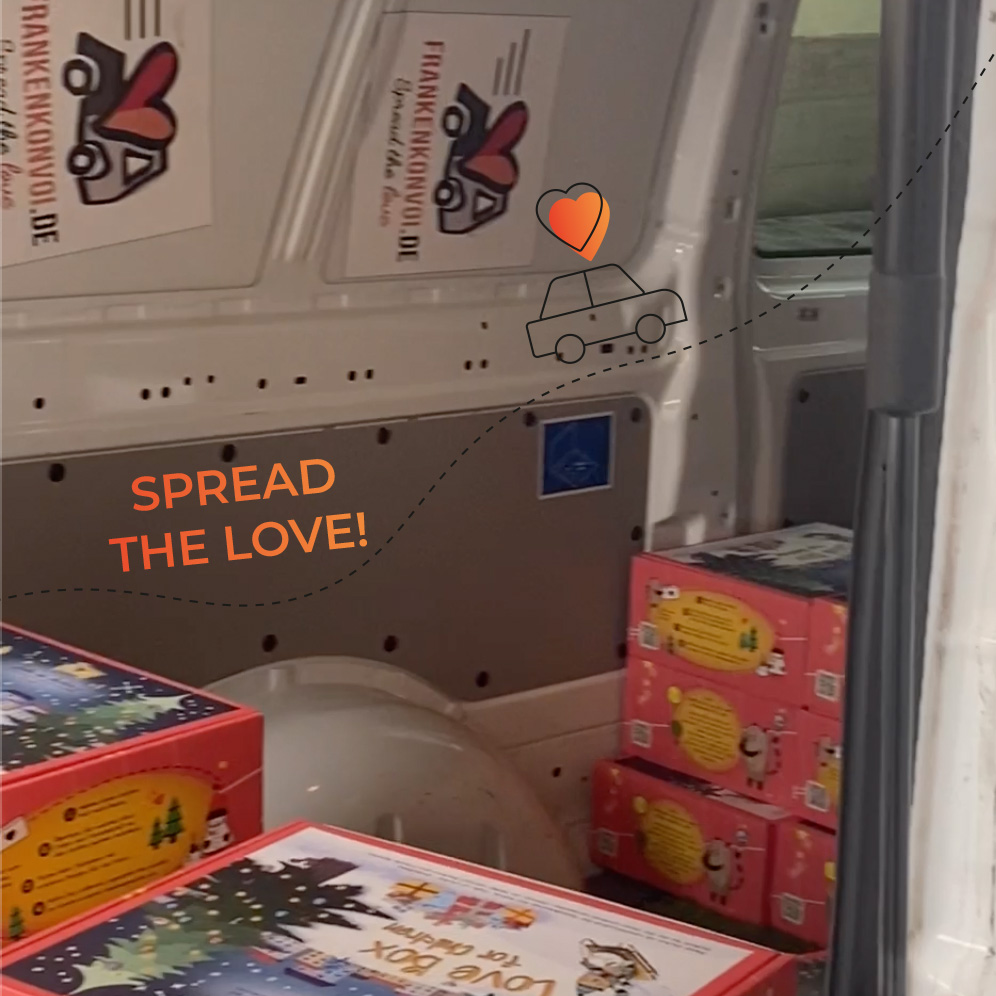 Im Inneren eines Transporters stapeln sich neben der offenen Tür mehrere bunte Geschenkboxen. An der Innenwand des Transporters sind Schilder mit einer Herz- und Auto-Illustration und dem Text "Spread the Love!" zu sehen