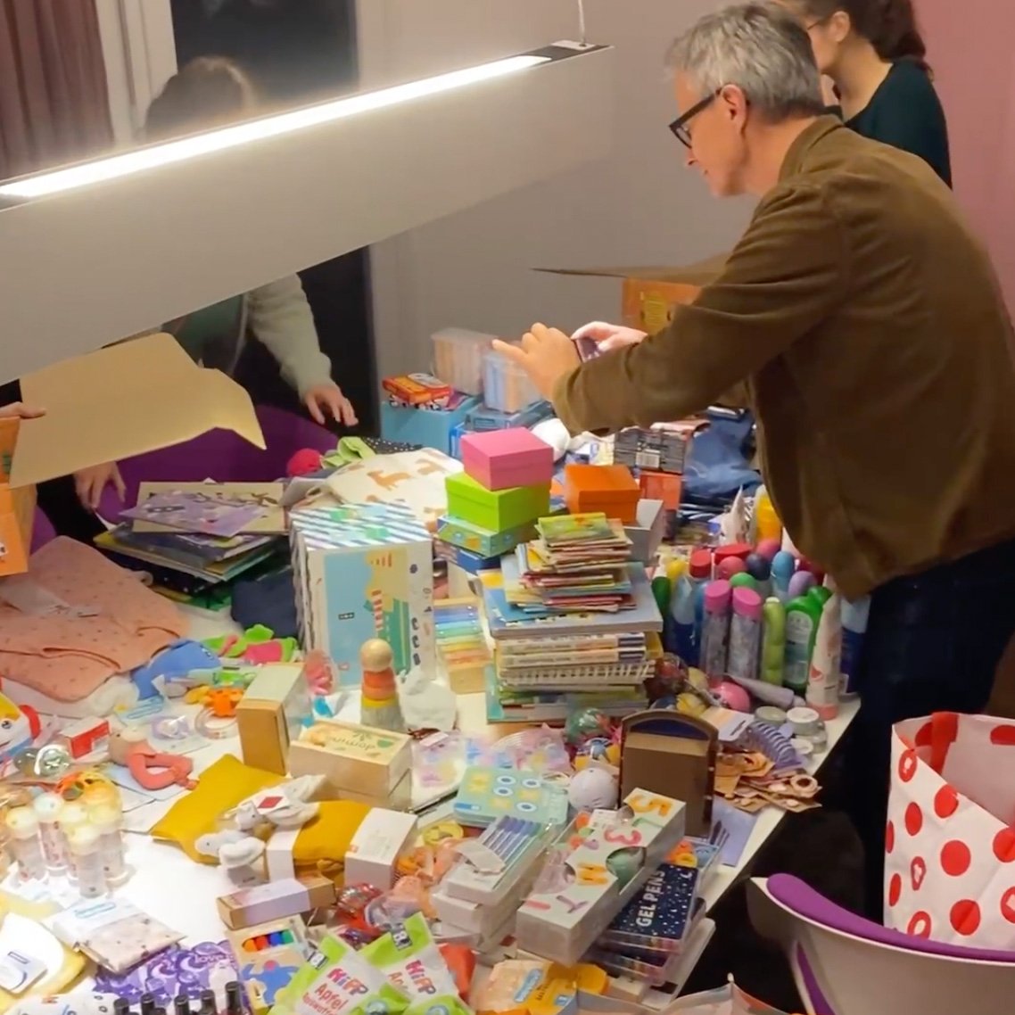 Zwei Personen organisieren einen Tisch voller bunter Gegenstände, von Büchern über Kunsthandwerk bis hin zu Hygieneartikeln, unter einem hellen Licht in einem gemütlichen Innenraum