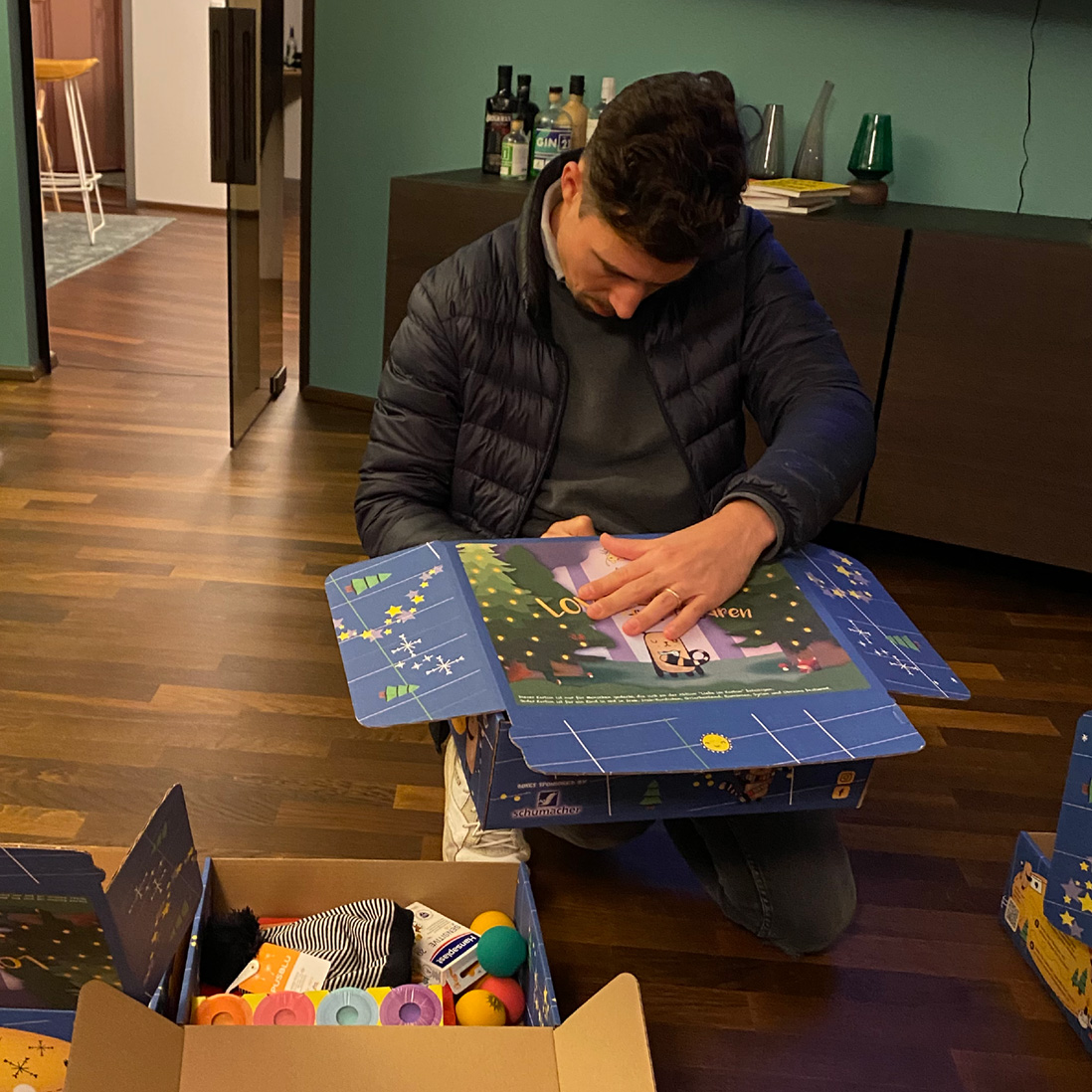 Ein Mann kniet auf einem Holzboden und öffnet eine blaue, weihnachtlich gestaltete Schachtel. In der Nähe steht eine offene Schachtel mit buntem Spielzeug und Geschenken. Der Raum hat eine tealfarbene Wand, Schränke und Flaschen auf einem Tresen