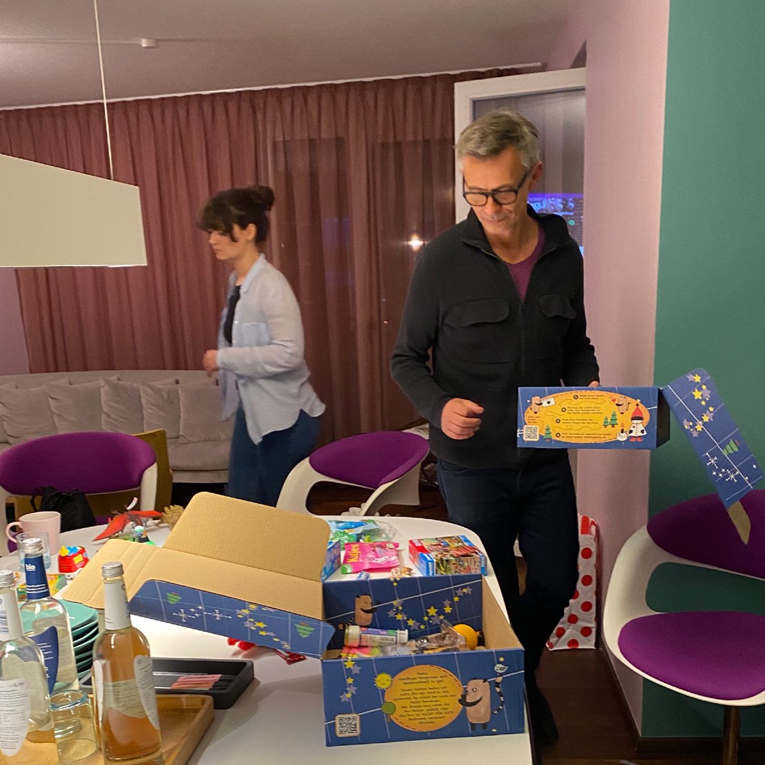 Ein Mann und eine Frau packen in einem Wohnzimmer bunte Geschenkkartons auf einem Tisch aus, der mit Flaschen, Geschenken und Snacks vollgestellt ist. Der gemütliche Raum mit lila Stühlen und rosa Vorhängen spiegelt die einladende Atmosphäre wider.