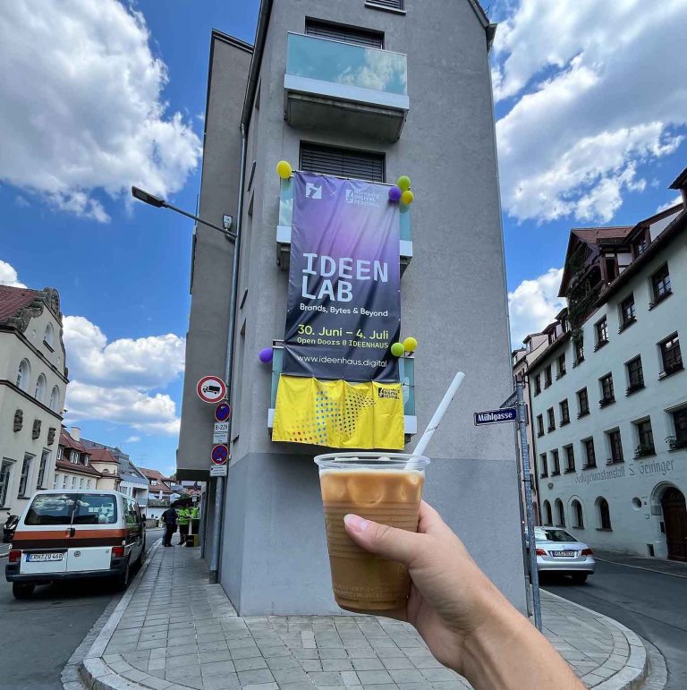 Eine Hand hält einen Eiskaffee mit einem Strohhalm im Vordergrund. Im Hintergrund zeigt ein hohes Gebäude ein Banner für "IDEEN LAB" - in der Nähe schweben bunte Luftballons, die unter einem strahlend blauen Himmel mit vereinzelten Wolken feiern, dass Wissen eine Marke ist.