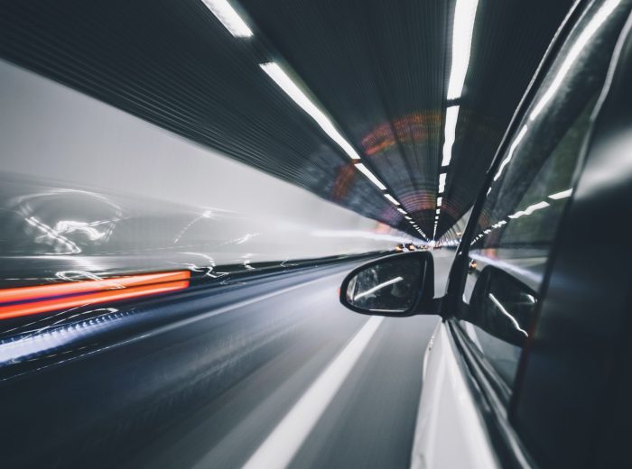 Ein weißes Auto rast durch einen nächtlichen Tunnel, aufgenommen von der Beifahrerseite. Markendehnung ist offensichtlich, da verschwommene Lichter und Bewegungsstreifen das Gefühl von schneller Bewegung und dynamischer Energie unterstreichen.