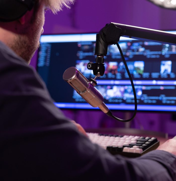 Eine Person mit Kopfhörern spricht in ein Mikrofon vor einem Computermonitor, auf dem mehrere Video-Miniaturansichten angezeigt werden, was auf ein Live-Streaming- oder Podcasting-Setup mit Schwerpunkt auf der Positionierung von Arbeitern hindeutet.